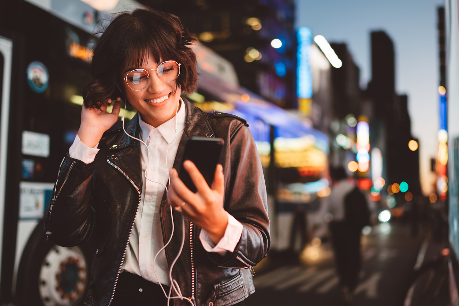 Iloinen silmälasipäinen nainen kuuntelee musiikkia kuulokkeilla kännykästä kävellessään kaupungin valaistussa keskustassa.